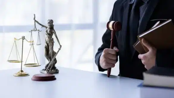 Un juge en costume noir tenant un marteau de justice et des livres de droit, avec une balance dorée et une statue de la justice sur un bureau blanc.